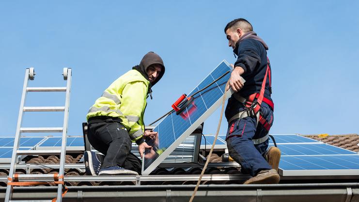 Zwei Arbeiter installieren ein Solarmodul auf einem Dach, während eine Leiter im Hintergrund steht.