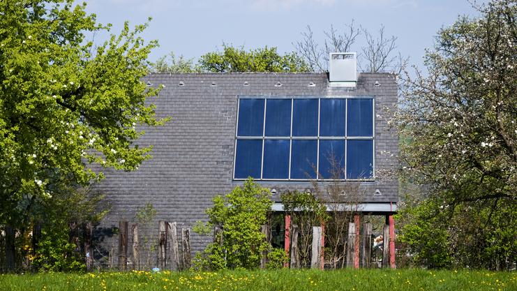 Moderne, umweltfreundliche Wohnhausfassade mit Solarzellen, umgeben von grünen Bäumen und blühenden Pflanzen im Vordergrund.